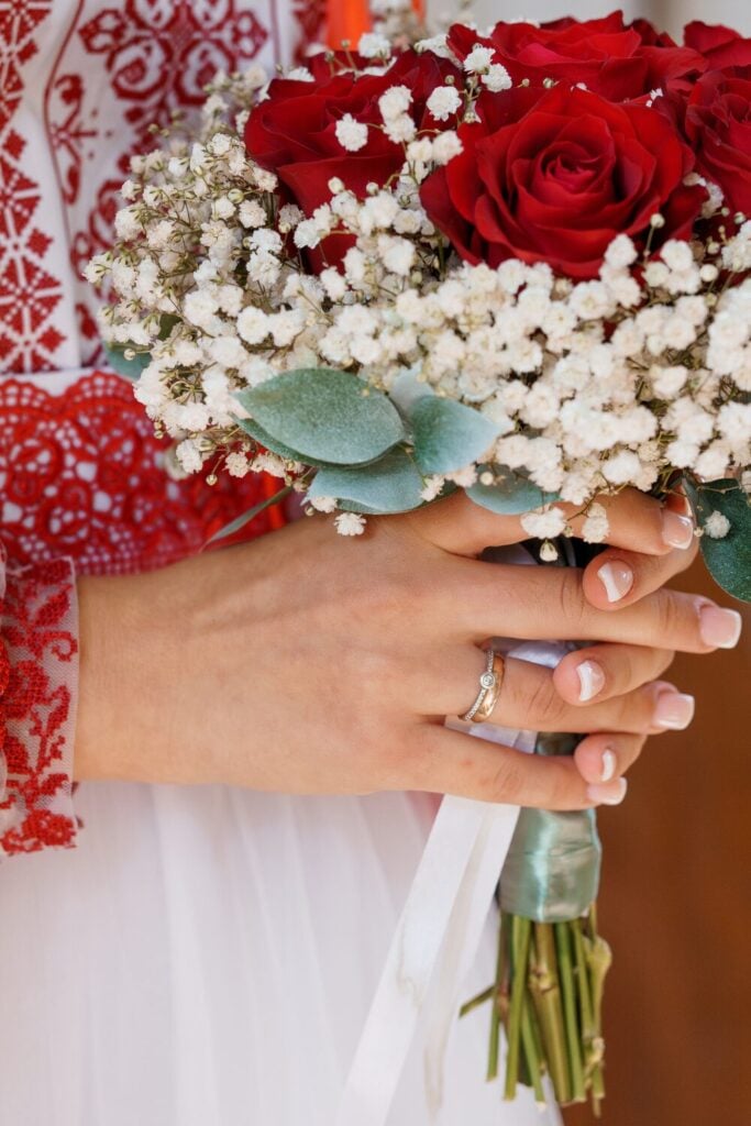 Classic red rose bouquet accented with Baby’s Breath and soft greenery, held by a bride with an elegant white and red embroidered dress.
