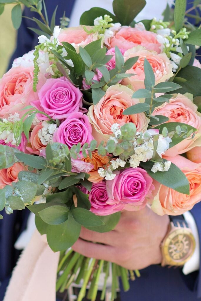 Vibrant bouquet of peach garden roses, pink blooms, fresh greenery, and Baby’s Breath, held by a groom in a navy suit.