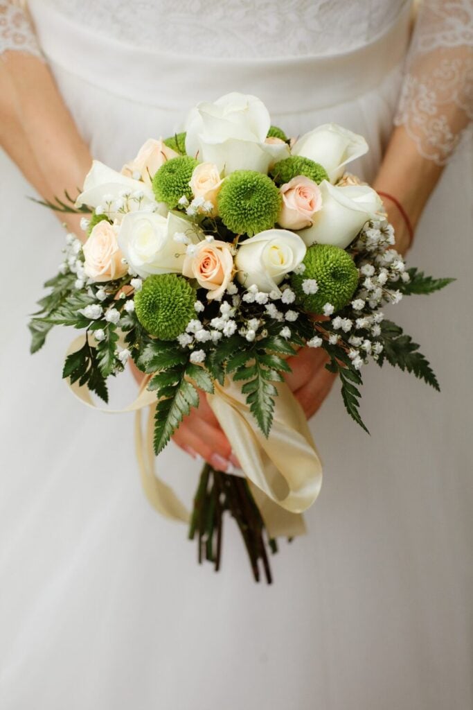 Bridal bouquet with white and peach roses, green button mums, and Baby’s Breath, tied with a cream ribbon for a fresh and elegant look.