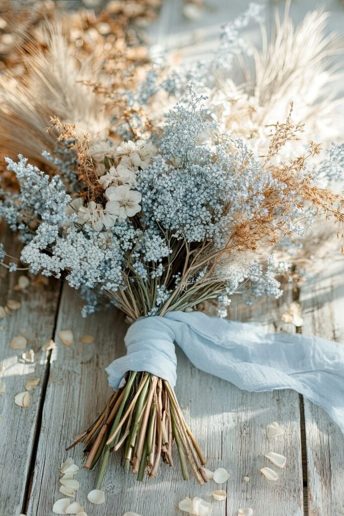 Rustic wedding bouquet with blue Baby’s Breath, dried grasses, ivory blooms, and a white ribbon.
